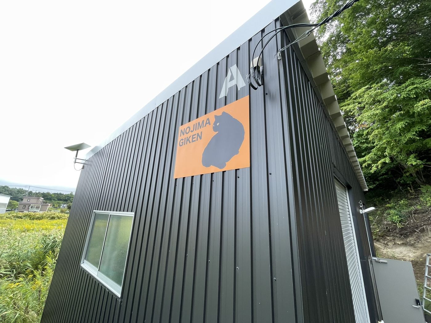 野島技研のガレージ工房の外観。黒い外壁に橙色の NOJIMA GIKEN 看板とシャッター